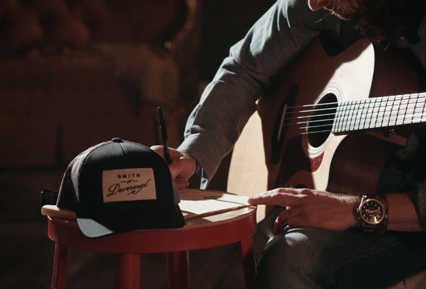 Close-up of a musician writing lyrics on a notepad while holding an acoustic guitar, with a Smith Devereux branded hat on a stool nearby.