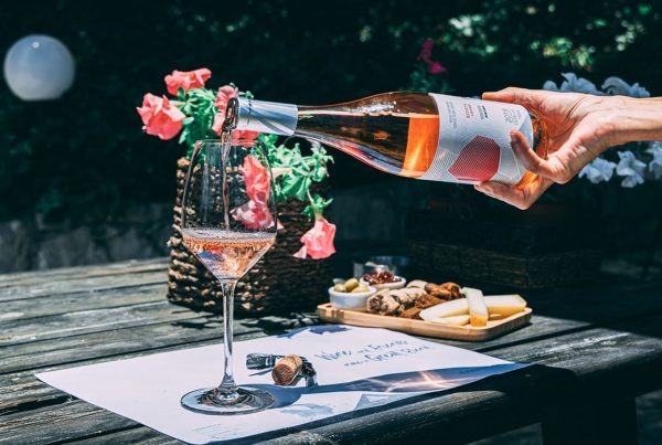 A hand pouring rosé wine from a bottle into a wine glass on a wooden table outdoors.