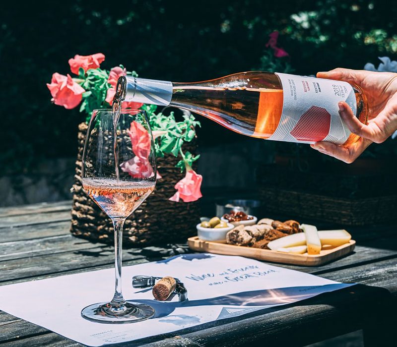 A hand pouring rosé wine from a bottle into a wine glass on a wooden table outdoors.