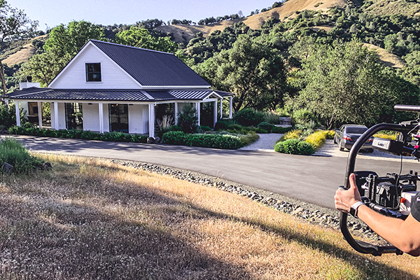 A person is holding a camera rig, filming a white house with a black roof in a scenic, hilly area with trees and greenery.
