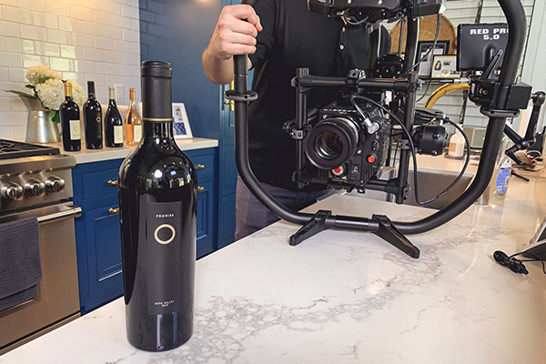 A person is holding a camera rig on a kitchen counter. In the foreground, there is a bottle of wine.