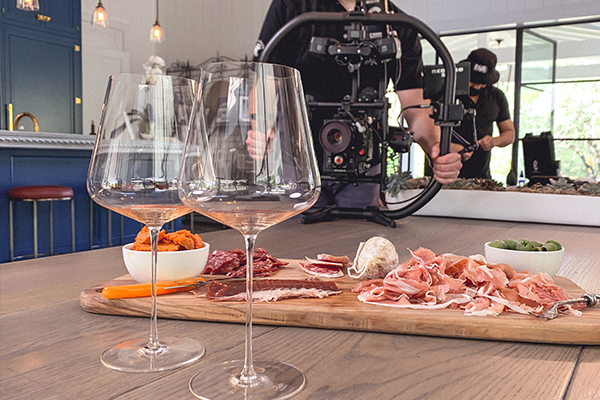 A close-up of a charcuterie board along with two wine glasses on a wooden table. In the background, a person operates a camera rig.