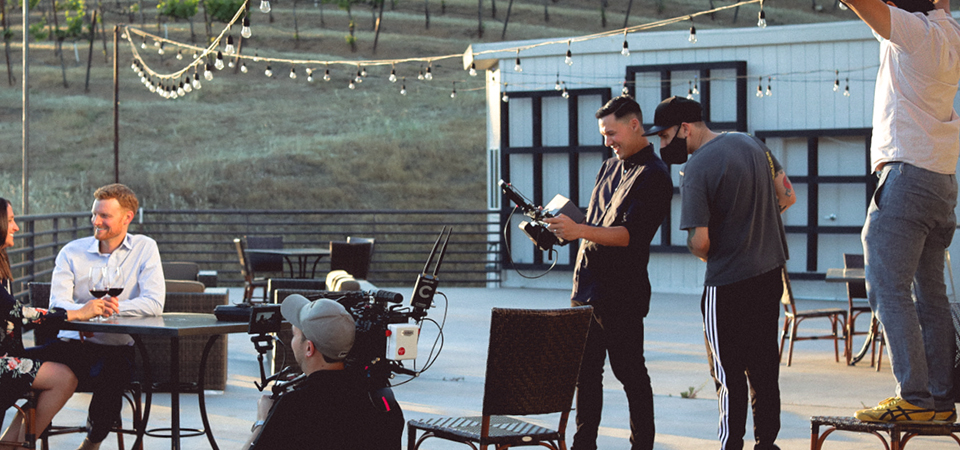 BTS shoot. Two people are seated at a table, engaged in conversation. In the foreground, there is a person operating a professional video camera.