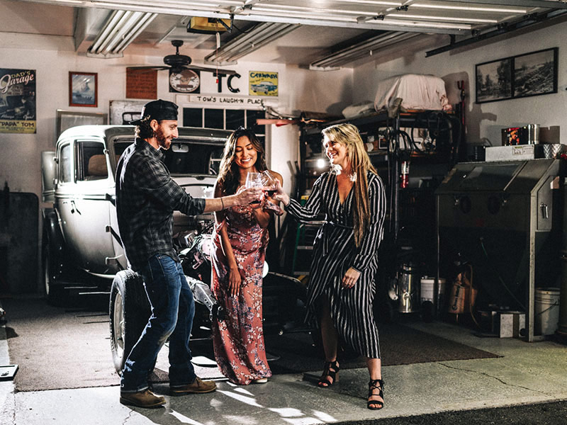 Three people standing in a garage with a vintage car in the background, sharing a drink.