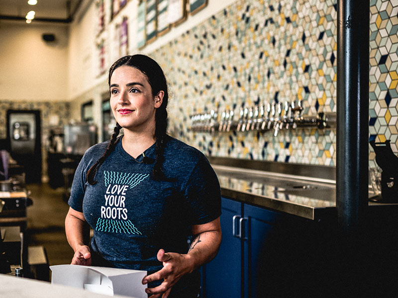 A woman in a bar with shelves of liquor bottles behind her.