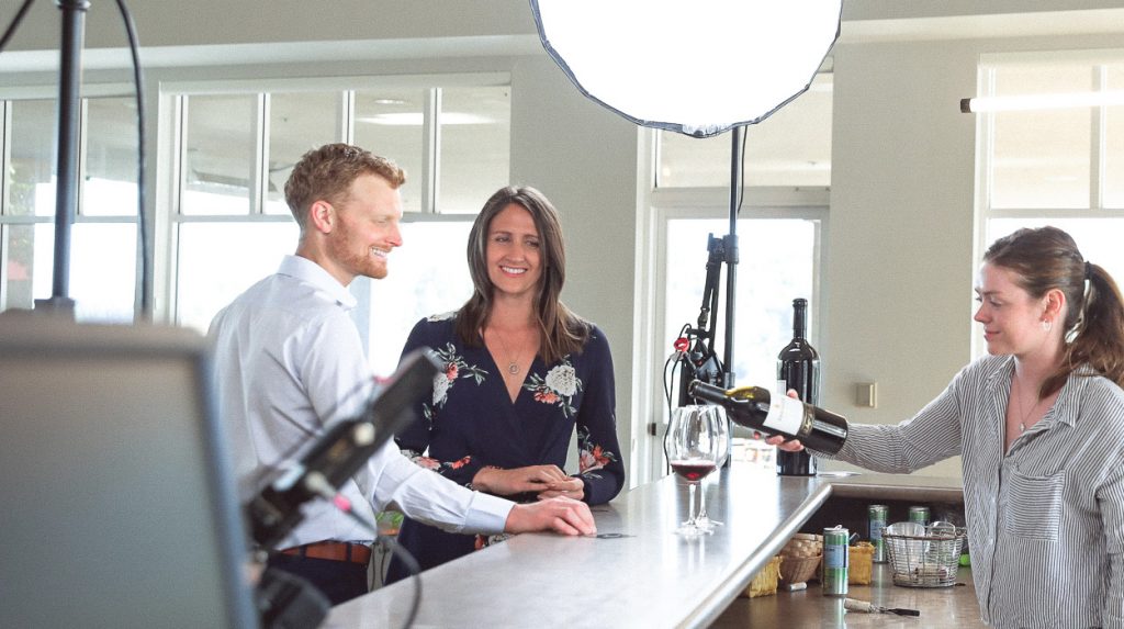 WineryHunt Case Study Still Banner Virtual A person is pouring red wine into a glass for another person at a bar counter in a production set.