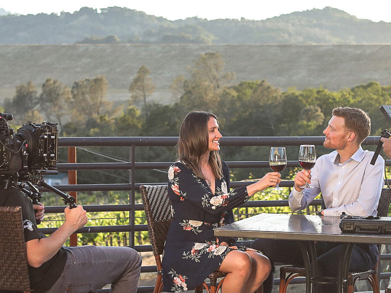 WineryHunt Case Study Still 1 A camera crew is filming two people sitting at an outdoor table on a balcony, toasting with glasses of red wine.