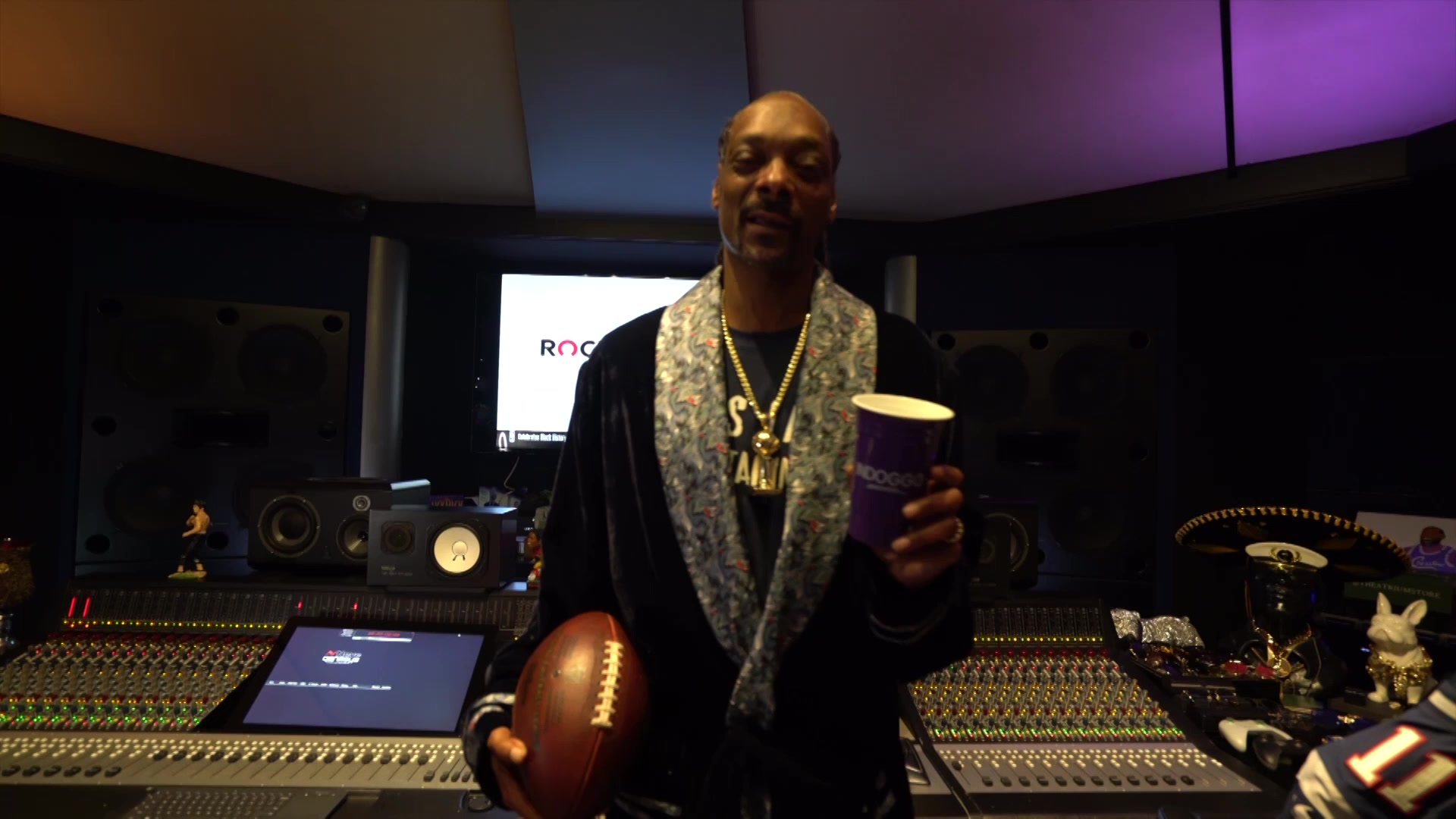 Snoop Dogg holding a football and a purple cup in a recording studio with a mixing console in the background.