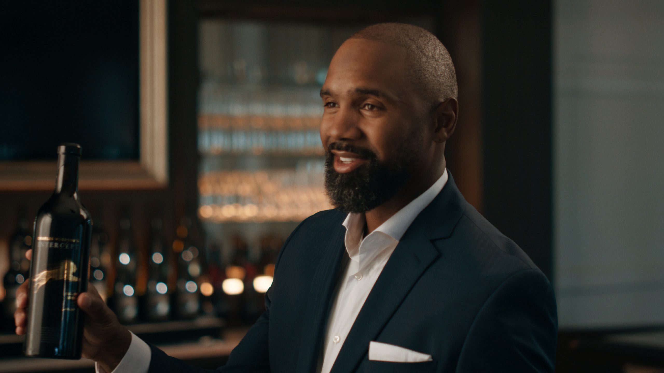 Charles Woodson in a suit holding a bottle of wine in a dimly lit bar area.