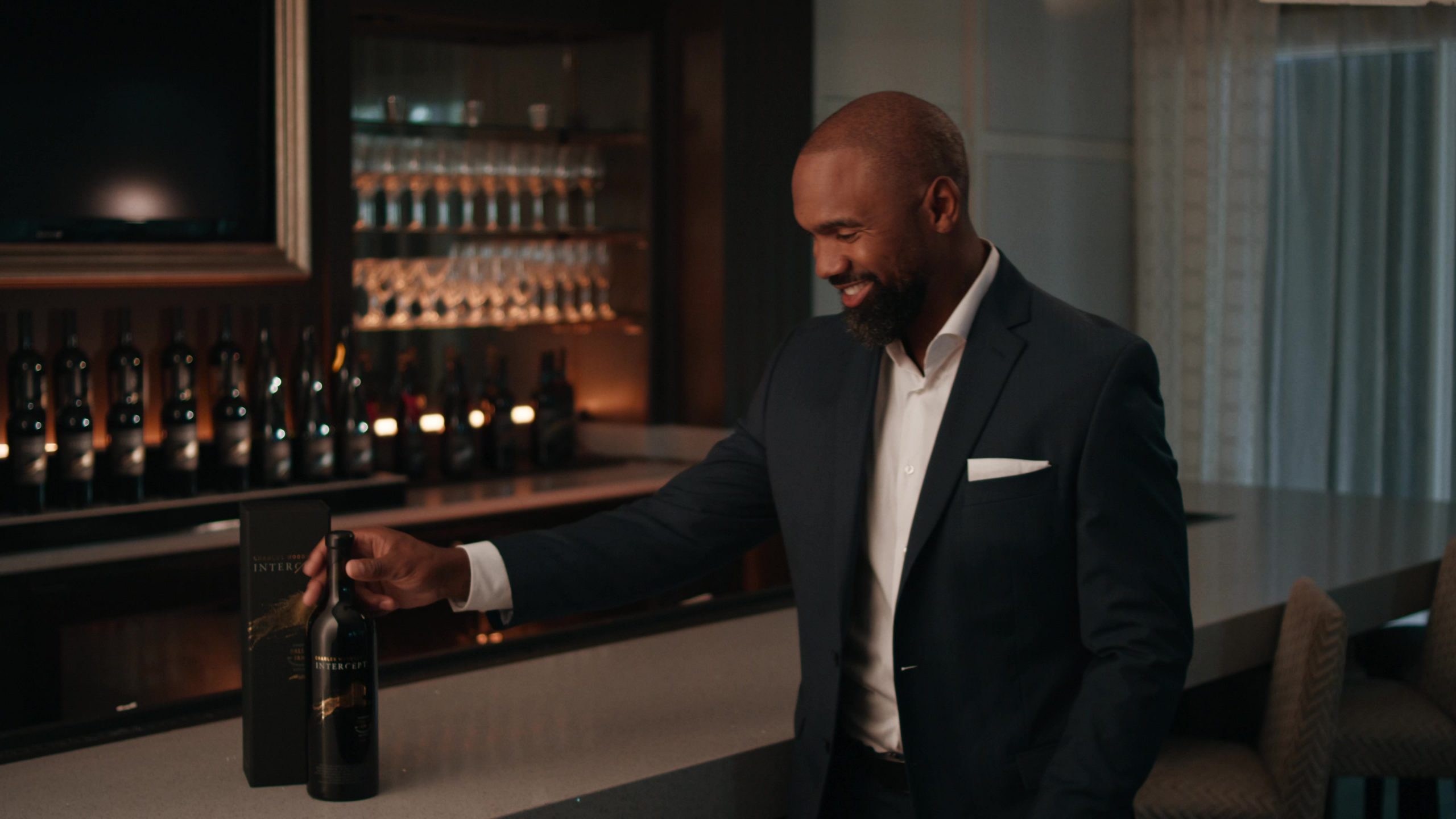 Charles Woodson in a suit holding a bottle of wine and looking at it in a dimly lit bar area.