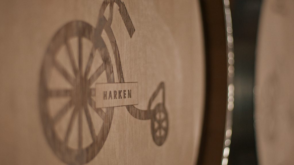 Close-up of a wooden barrel with a logo of a penny-farthing bicycle etched onto its surface, labeled HARKEN.