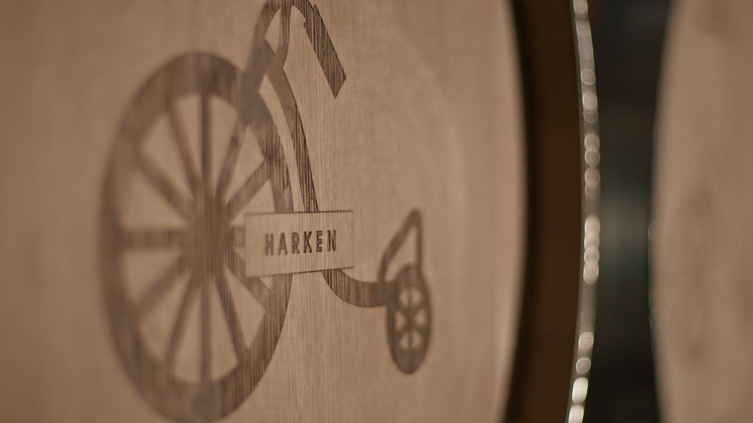 Close-up of a wooden barrel with a logo of a penny-farthing bicycle etched onto its surface, labeled HARKEN.