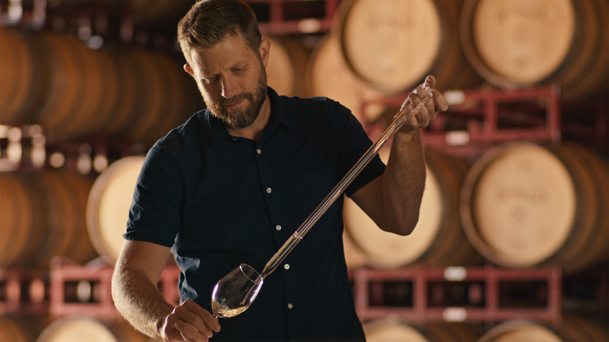 A man holding a wine thief in a wine cellar with wooden barrels.