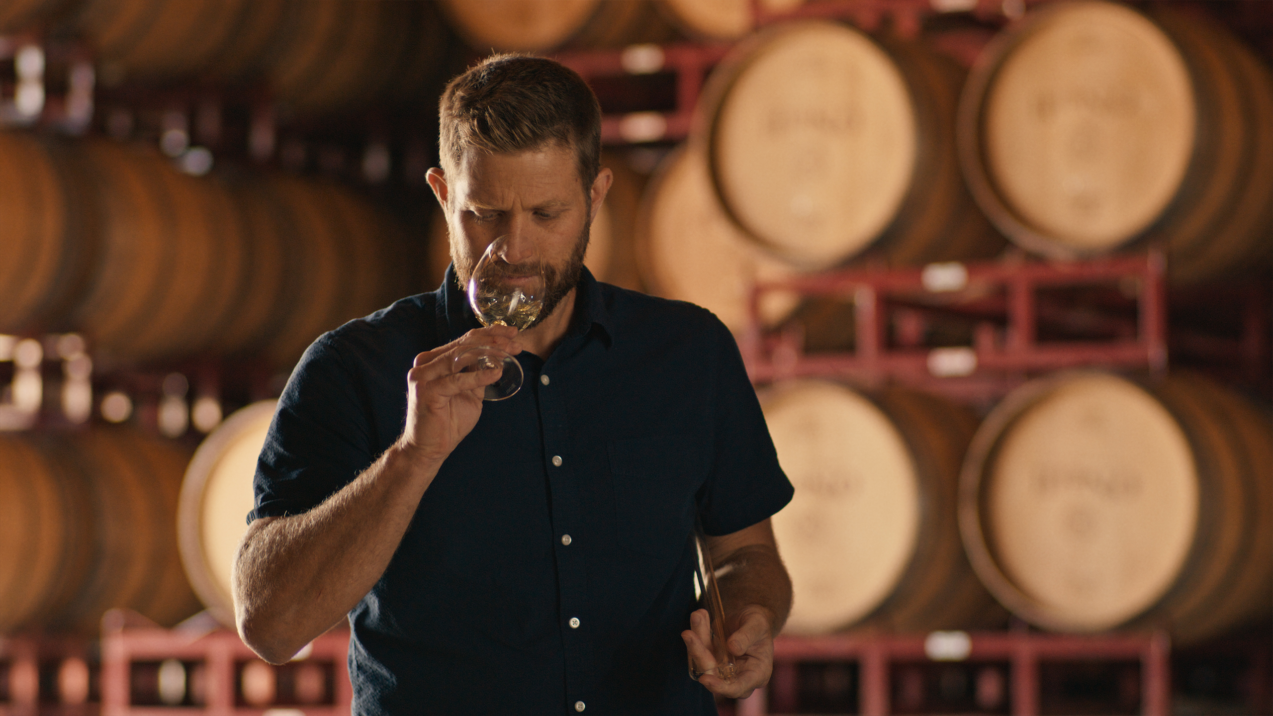 A man in a navy blue shirt tasting wine in a dimly lit winery, surrounded by wooden barrels stacked on racks.