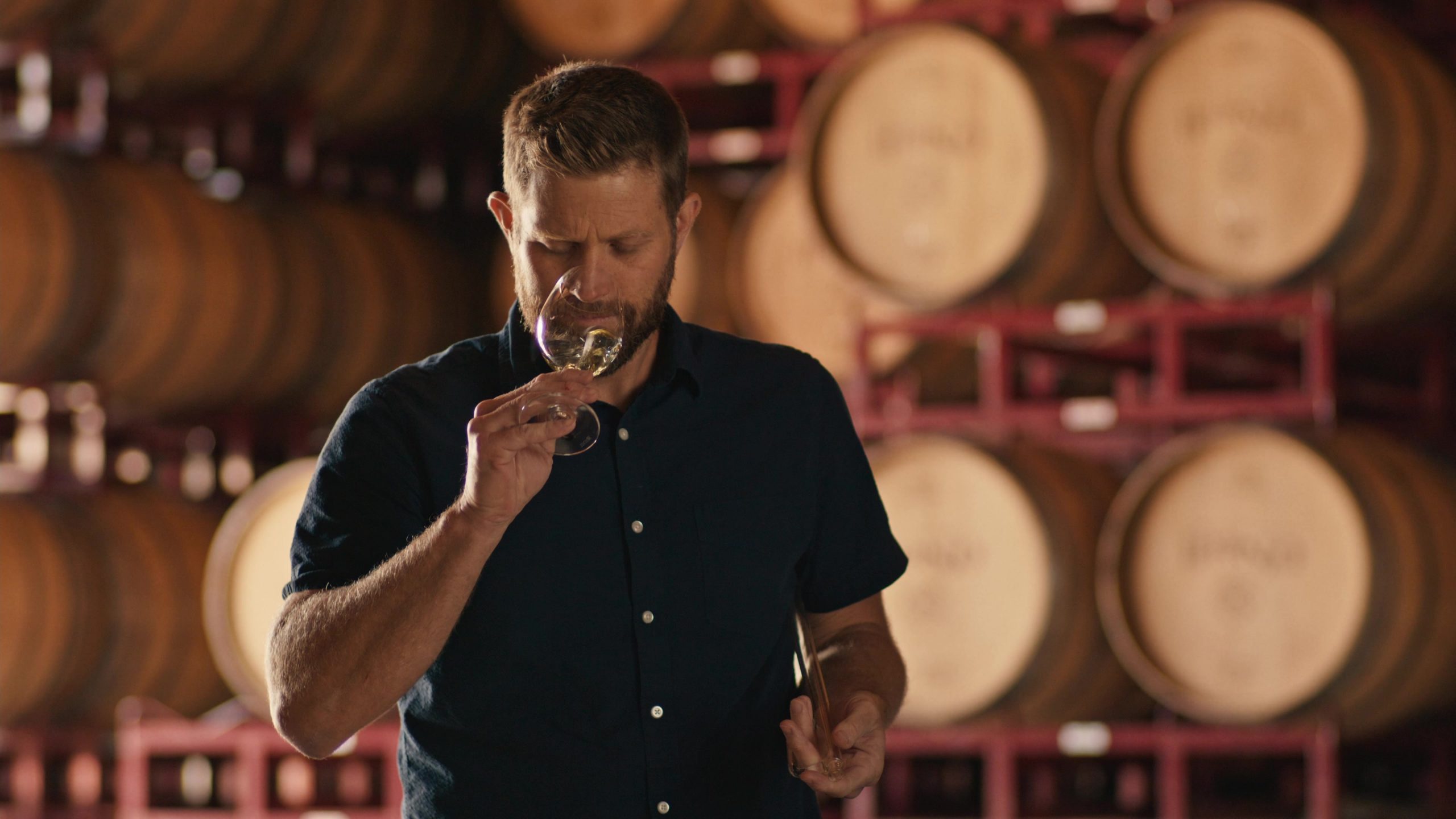 A man holding a wine glass and clipboard in front of stacked wooden barrels in a winery or distillery.