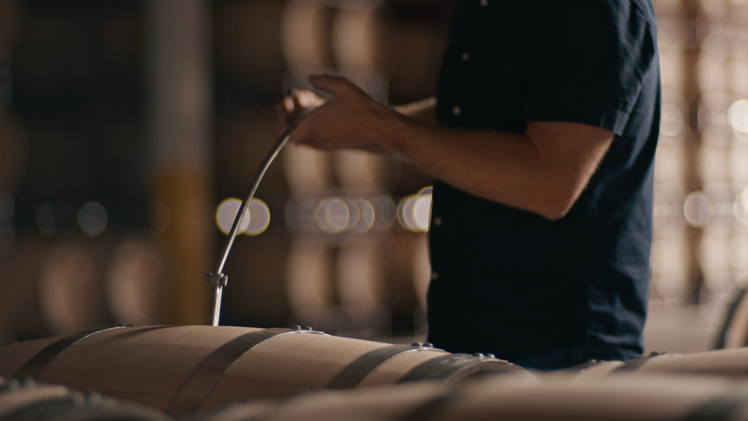 Person using a tool to extract liquid from a barrel in a storage facility with multiple barrels in the background.