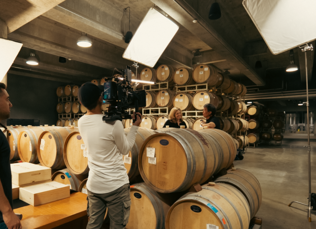A wine cellar with wooden barrels in the background. A man films two people sitting and talking near the barrels.