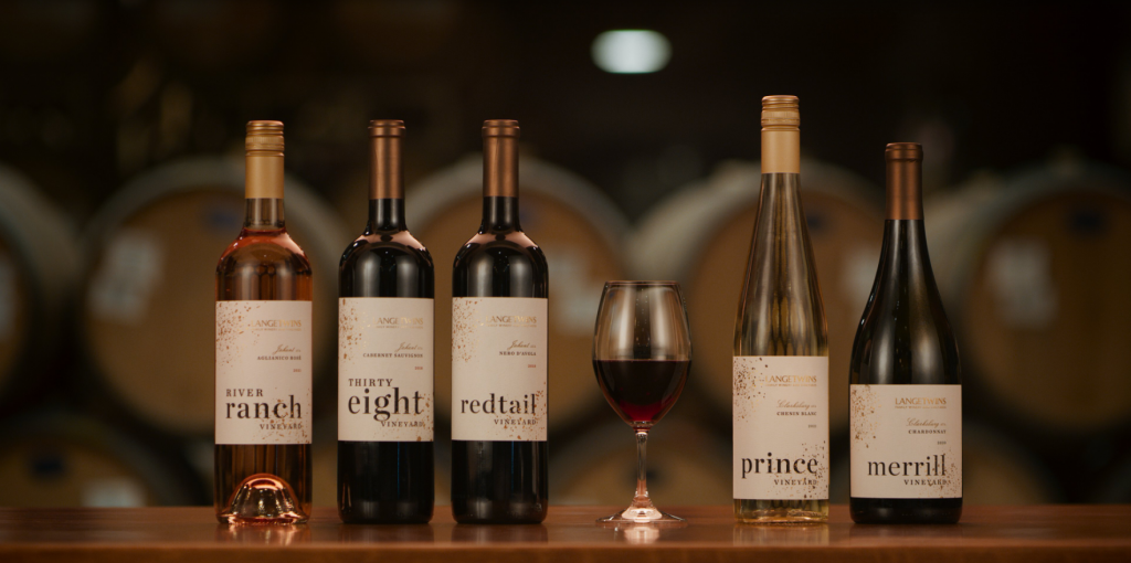 Five bottles of wine labeled 'River Ranch,' 'Thirty Eight,' 'Redtail,' 'Prince,' and 'Merrill,' with a glass of red wine between the third and fourth bottles, set on a wooden surface.