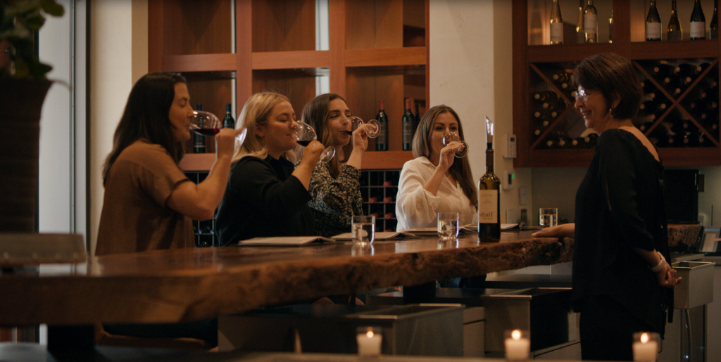 Group of four women seated at a bar counter, each holding and tasting wine glasses, with a fifth woman guiding the session.