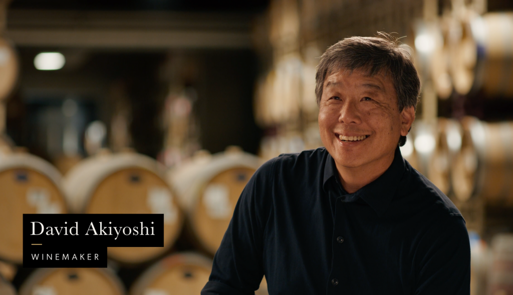 A man standing in a wine cellar with wine barrels in the background and a label that reads 'David Akiyoshi, Winemaker.'